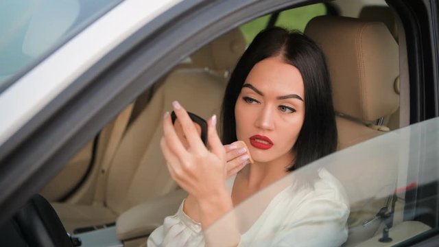 Beautiful elegant brunette woman with red lipstick on her lips applying makeup powder in her expensive car decorated with white leather interior