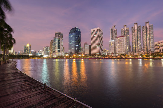 Scenic view of wooden boardwalk and lake at the Benjakiti (Benjakitti) Park and lit skyscrapers in Bangkok, Thailand, at sunset.
