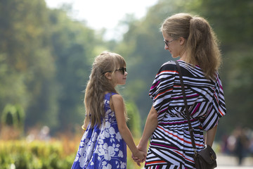 Back view of young blond long-haired attractive woman and small child girl holding hands outdoors.