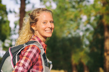 Hiking blonde woman healthy and active walking with backpack smiling happy in forest. Female hiker backpacker trekking in summer outdoors. Beautiful young adult girl living a happy lifestyle.