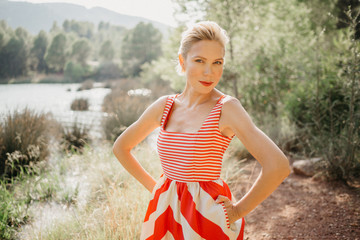 A stylish blonde lady in the red and white dress posing near the swamp of the mountain lake. Forward look. Close portrait. Spain