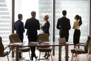 Multiracial workers colleagues talk at break, drink coffee, stand near window, modern city skyscrapers. Coworkers in office discuss future project, planning new opportunities. Business vision concept