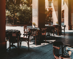 Fototapeta na wymiar Cozy outdoor cafe with empty wooden tables covered with black tablecloths, metal flower pots and ashtries on them, chairs near; exterior of an empty street bar on the daytime, pavement below