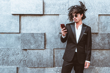 Curly stylish Asian guy in the sunglasses and in a formal suit is standing outdoors in front of...