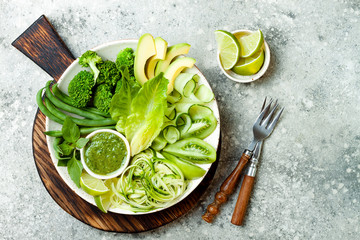 Vegan, detox Buddha bowl with avocado, zucchini noodles, green beans, spiralized cucumber, tomatoes, broccoli, lime and pesto sauce. Top view, grey concrete background