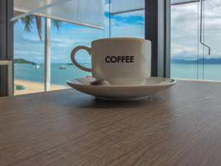 Cup of coffee placed on a wooden table with a tropical seascape in the background.