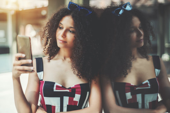 A charming curly caucasian girl is typing a message on her smartphone while leaning against glass wall; dazzling young Moroccan woman is using her cell phone while waiting for her friend outdoors - Powered by Adobe