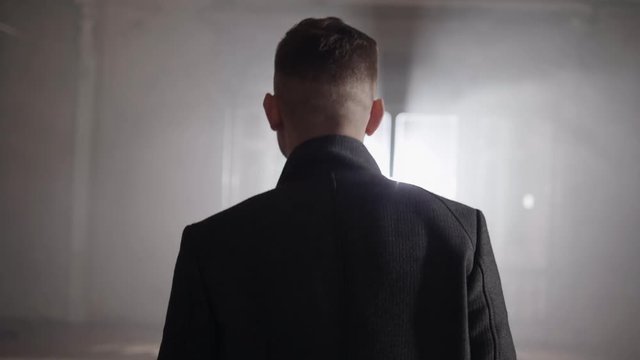 Short haired man wearing long grey jacket with straight collar is walking in darken empty hall towards bright lighting coming out of open windows