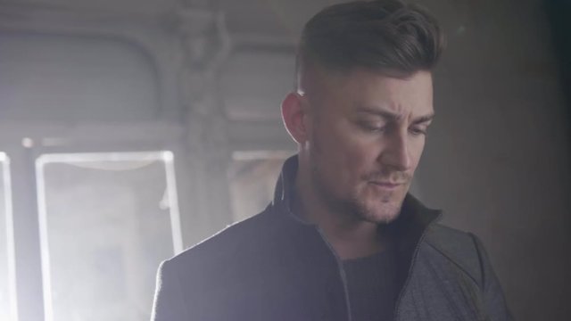 Handsome seriously looking bearded young man with grey eyes and stylish haircut dressed in classic darken coat with straight collar starts walking away from windows with bright lighting.