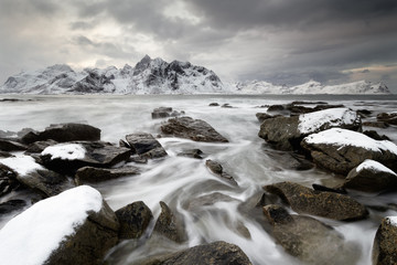 Fototapeta na wymiar Coastal landscape in winter with water movement between big stones, in the background a mountain range with snow, high contrast sky - Location: Norway, Lofoten