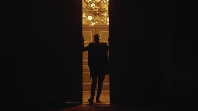 Young slim man opens two large massive doors and enters into darken empty hall leaving behind room with big chandalier consisting of several yellow bulb looking lamps mounted to ceiling.