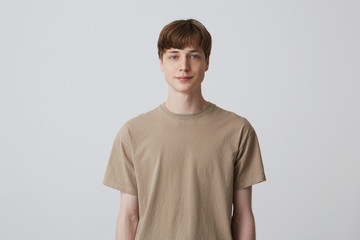 Portrait of handsome attractive young man with short haircut and blue eyes wears beige t shirt standing and smiling isolated over white background Looks directly in camera