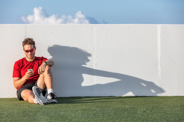 Fitness sport man using mobile phone app after workout training relaxing sitting on outdoor grass post run.