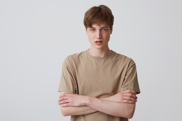 Portrait of upset unhappy young man with short haircut in beige t shirt looks desperate and standing with arms crossed isolated over white background © timtimphoto