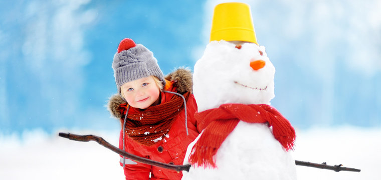 Little boy in red winter clothes having fun with snowman in snowy park