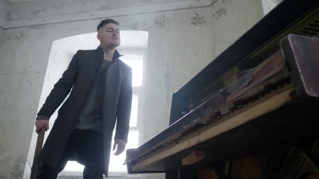 Handsome young bearded man with modern short haircut dressed in grey coat is agressively smashing abandoned classic wooden piano with huge metallic hammer in cold room.