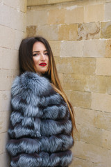 Young attractive girl in a fur coat in a winter park. She stands by the brick wall. Street fashion.