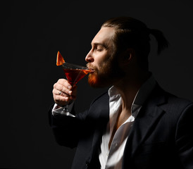 Young business man in white shirt and jacket drinking red martini cocktail drink  looking at the corner on dark 