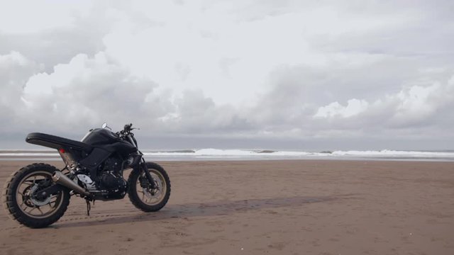 Modern custom motorcycle cafe racer on the black sand ocean beach near the water. Adventure and travel concept. Surfing spot with ocean waves.