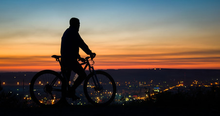 Obraz premium A young cyclist rests on the top of the mountain near the city, at sunset.