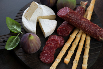 Close-up of sliced salami, camembert cheese, grissini and ripe fig fruits on a black wooden serving board