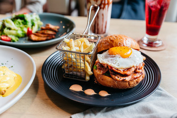 Tasty burger with fried egg served with fries in black plate on wooden table.