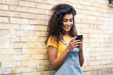 Happy Arab girl using smart phone on brick wall.