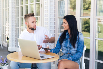 An attractive guy with a stylish haircut and a beard tells something to a brunette European. A girl listens attentively to a man, holds a smartphone in her hand, works on a laptop. Concept of people