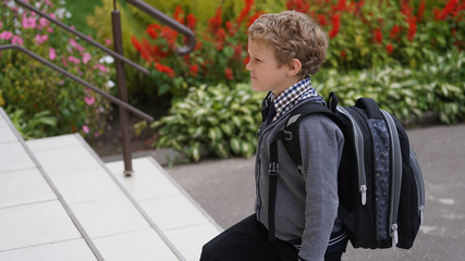Little Caucasian curly boy in school uniform with backpack comes up on stairs.