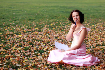 Fine emotional brunette writing on paper, sitting on autumn leaves and grass. Copy space. Yonge girl in vintage pink pastel with live expressions on beautiful country and old background.