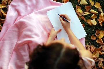 Fine emotional brunette writing on paper, sitting on autumn leaves and grass. Copy space. Yonge girl in vintage pink pastel with live expressions on beautiful country and old background.