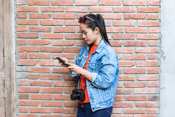 Woman with sunglasses on head, wearing a jacket, jeans and orange shirt standing against a wall and playing mobile phones with cameras.