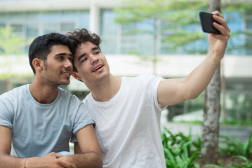 Happy interracial gays posing for cute selfie in city. Cheerful young friends in t-shirts photographing together against university building. Relationship concept 