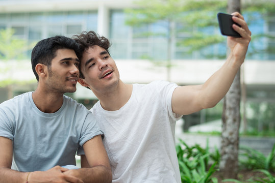 Happy interracial gays posing for cute selfie in city. Cheerful young friends in t-shirts photographing together against university building. Relationship concept 