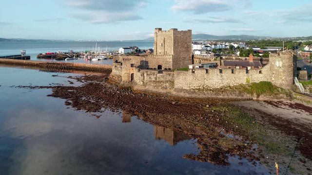 Medieval Norman Castle in Carrickfergus near Belfast in sunrise light. Aerial 4K flyby video with water reflection, marina, groin, breakwater,  town and far view of Belfast in the background