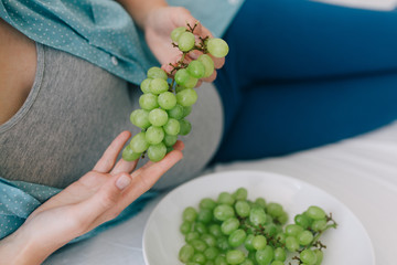 Cropped photo of young pregnant woman eating grapes in bed .