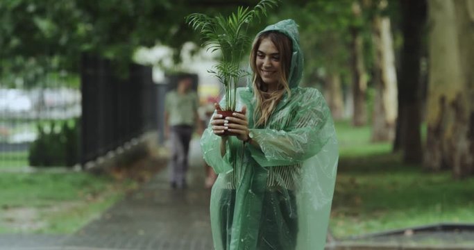 Smiling young woman holding in a rain day a flower in a pot.