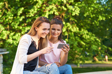 Two young girls are sitting in the Park on the background of trees and watching photos on smartphone. The concept of mobile devices, gadgets, wireless communication. Lifestyle and outdoor recreation