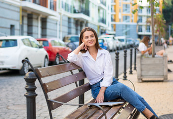 Young beautiful smiling female with a laptop sits on a bench in the business part of the city.