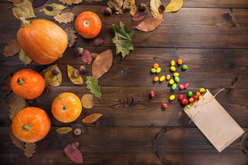 Happy Halloween! The concept of the autumn festival, small orange pumpkins with dry leaves lie on a brown wooden background, next to the scattered colored candy from a paper bag, top view