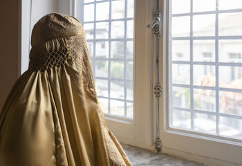 Indoor head and shoulder shot of a person wearing a gold-coloured Burqa looking through a grilled window to the outside