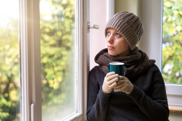 Frau steht an einem Fenster und trinkt ein heißes Getränk, Winter und aufwärmen