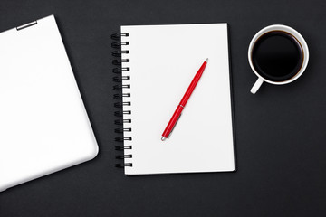 Top view workspace mockup on black background with blank notepad, red pen, coffee cup, headphones and closed silver laptop. Note pad space for text input. 