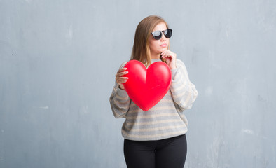 Young adult woman over grey grunge wall holding red heart in love serious face thinking about question, very confused idea