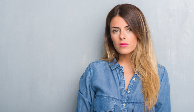 Young adult woman over grunge grey wall wearing denim outfit with serious expression on face. Simple and natural looking at the camera.