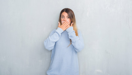 Young adult woman over grey grunge wall wearing winter outfit shocked covering mouth with hands for mistake. Secret concept.