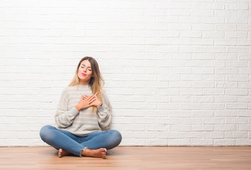 Young adult woman sitting on the floor over white brick wall at home smiling with hands on chest with closed eyes and grateful gesture on face. Health concept.