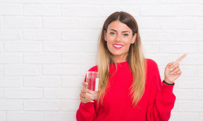 Young adult woman over white brick wall drinking glass of water very happy pointing with hand and finger to the side