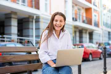 Young beautiful laughing female with a laptop sits on a bench in the business part of the city.