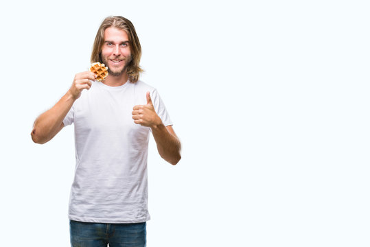 Young handsome man with long hair over isolated background eating waffle happy with big smile doing ok sign, thumb up with fingers, excellent sign